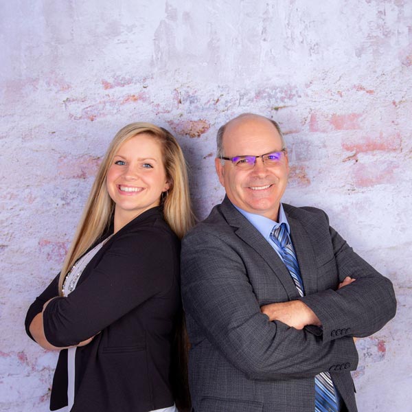 Hank and Laura standing back to back with their arms crossed in front of a white brick wall